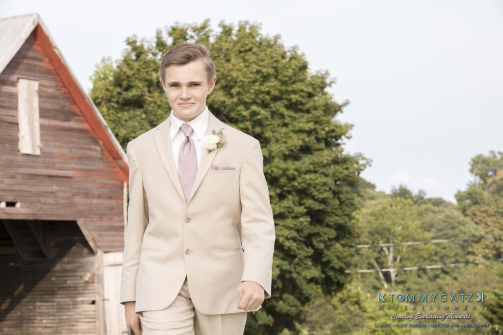 JenniferRudiWeddingCeremonyPhotos-74 - TOMMY GATZ ENTERTAINMENT A young man in a beige suit with a boutonniere walks outside near a wooden barn, with trees in the background.