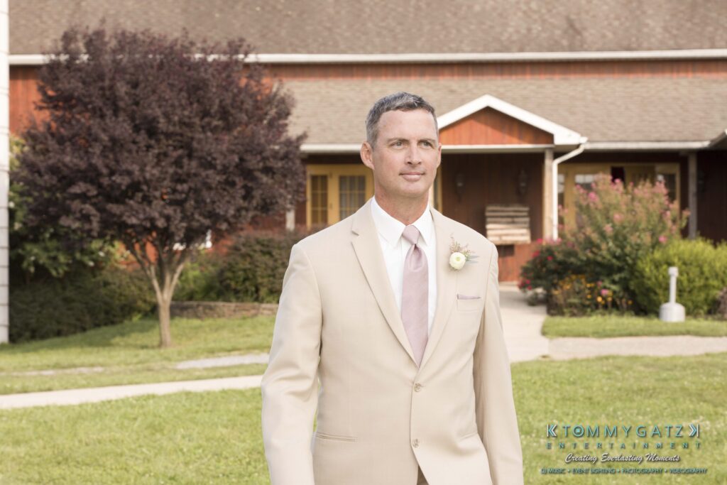 JenniferRudiWeddingCeremonyPhotos-75 - TOMMY GATZ ENTERTAINMENT Man in beige suit with pink tie and boutonniere stands on grass, with a brown building and tree in the background.