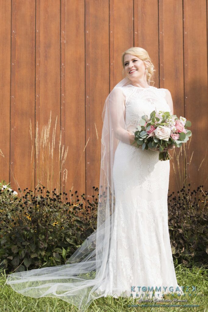JenniferRudiWeddingFormalPhotos-182 - TOMMY GATZ ENTERTAINMENT Bride in a white lace gown holds a bouquet of pink and white flowers, standing in front of a wooden wall with tall grass.