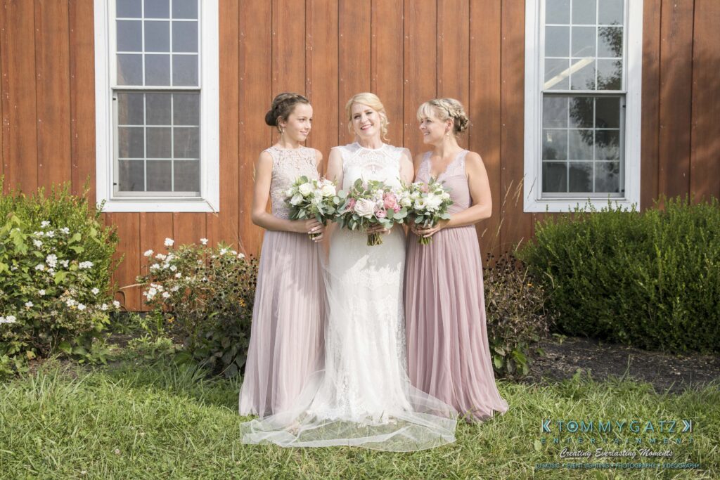 JenniferRudiWeddingFormalPhotos-184 - TOMMY GATZ ENTERTAINMENT Three women in pastel dresses stand together holding bouquets in front of a wooden building with two windows. The central woman wears a white dress. They are on a grassy area with shrubs.