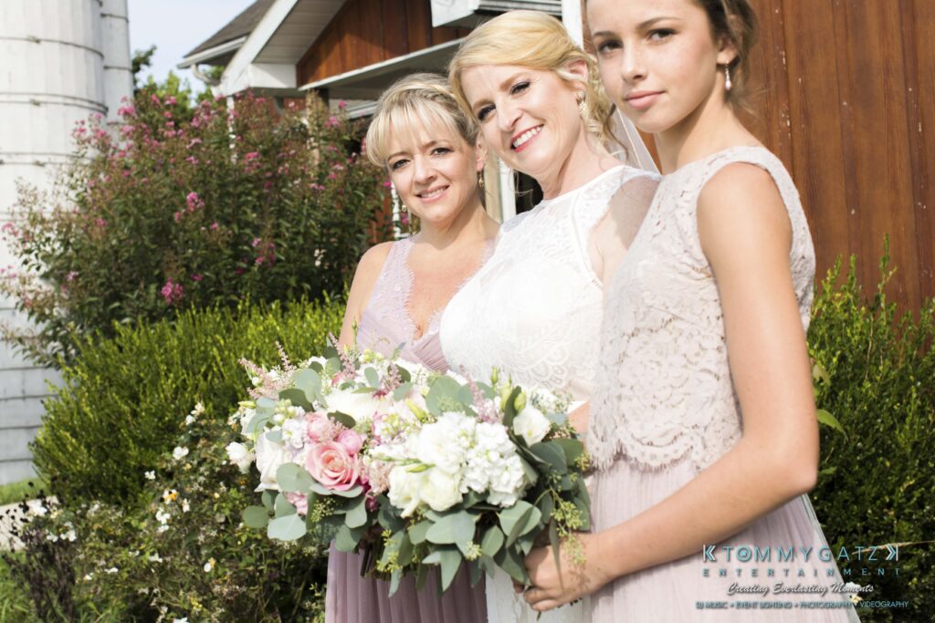 JenniferRudiWeddingFormalPhotos-196 - TOMMY GATZ ENTERTAINMENT Three women in pastel dresses pose outdoors. The woman in the center holds a bouquet of flowers. A house and silo are visible in the background.