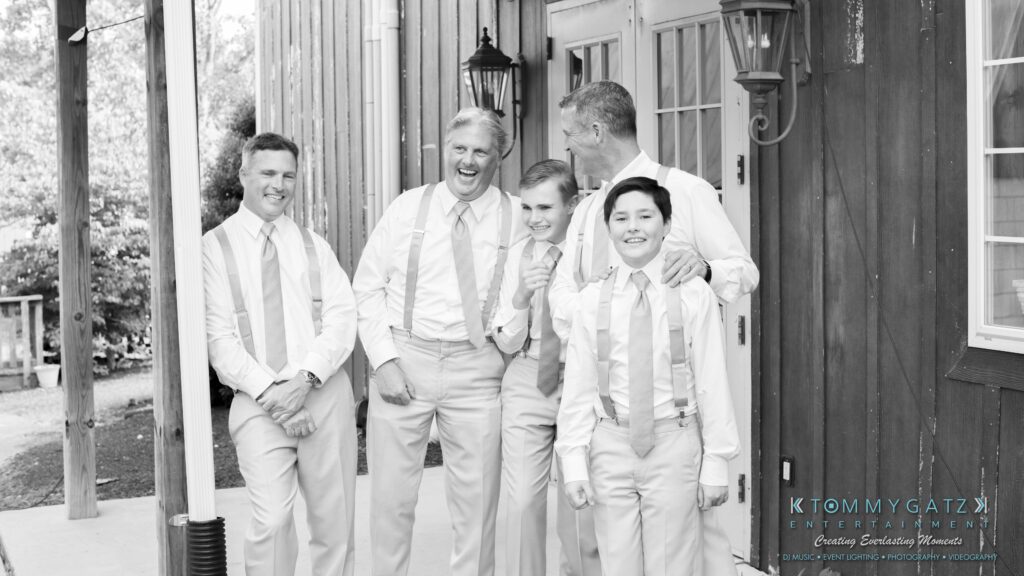 JenniferRudiWeddingFormalPhotos-201-1 - TOMMY GATZ ENTERTAINMENT Five men in dress shirts, ties, and suspenders stand laughing together outside a wooden building.