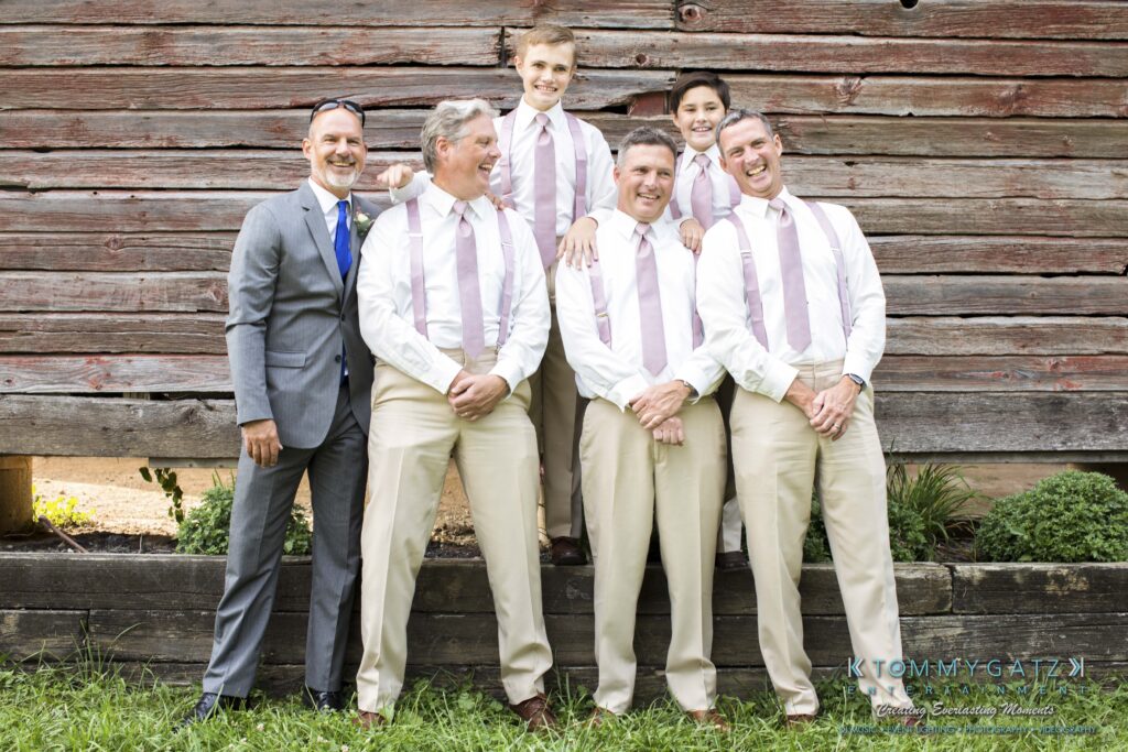 JenniferRudiWeddingFormalPhotos-204 - TOMMY GATZ ENTERTAINMENT Five men in formal attire pose in front of a rustic wooden wall, with four wearing matching suspenders and ties. They appear to be smiling.