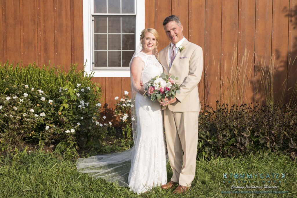 JenniferRudiWeddingFormalPhotos-209 - TOMMY GATZ ENTERTAINMENT A bride and groom stand smiling in front of a wooden building with a window. The bride holds a bouquet of flowers.