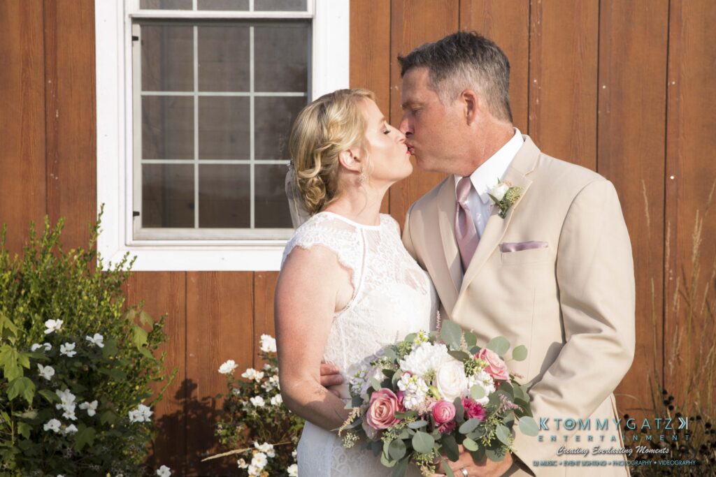 JenniferRudiWeddingFormalPhotos-210 - TOMMY GATZ ENTERTAINMENT A bride and groom share a kiss outdoors. The groom wears a beige suit, and the bride holds a bouquet of flowers. They stand in front of a wooden wall with a window.