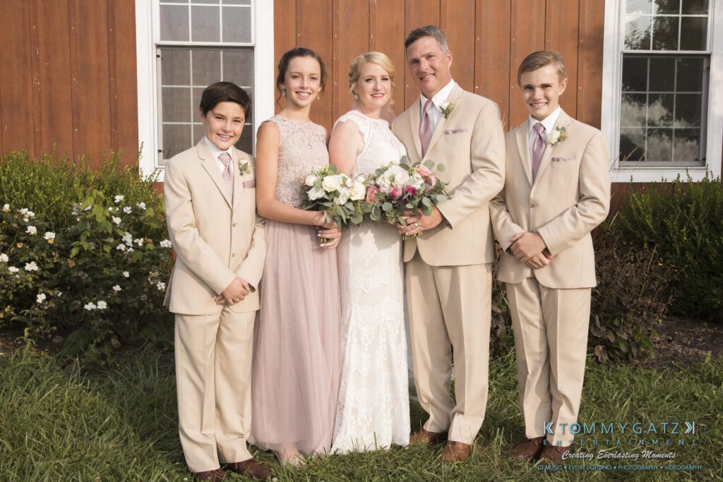JenniferRudiWeddingFormalPhotos-211 - TOMMY GATZ ENTERTAINMENT A family stands in front of a wooden building. Two young men in beige suits, a young woman and a bride in light dresses, and a man in a beige suit pose together, holding flowers.