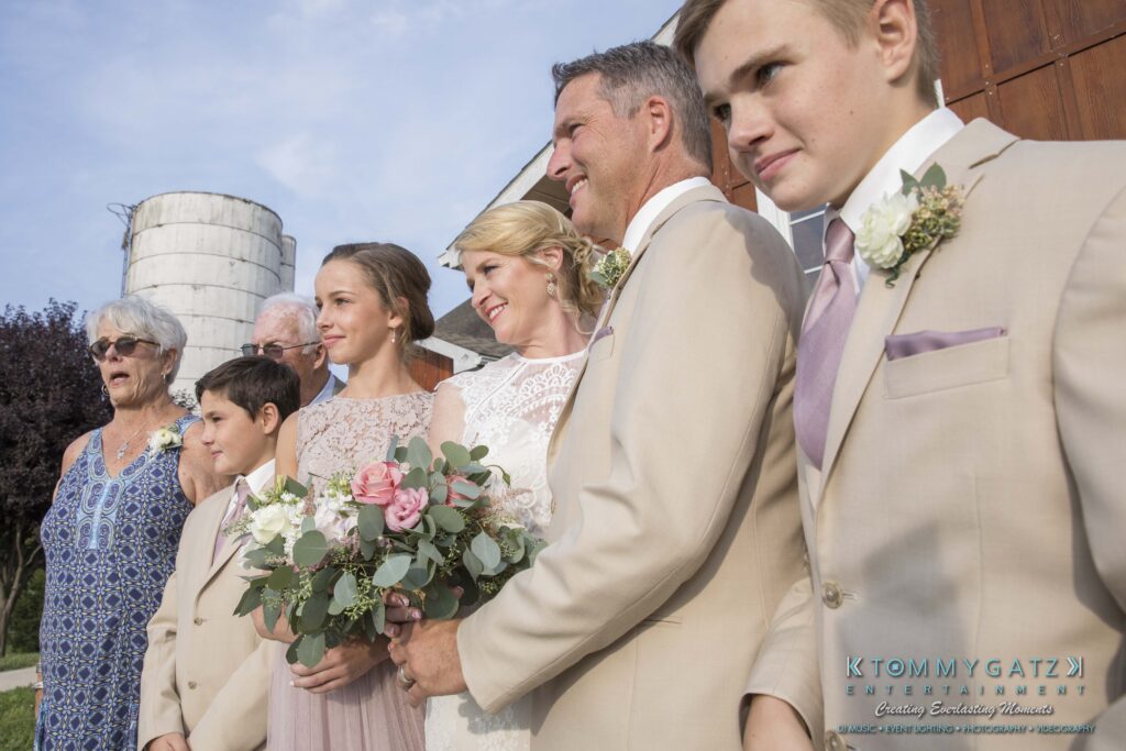 JenniferRudiWeddingFormalPhotos-212 - TOMMY GATZ ENTERTAINMENT A group of people in formal attire pose for a photo outdoors. A woman holds a bouquet. A silo and wooden structure are in the background.