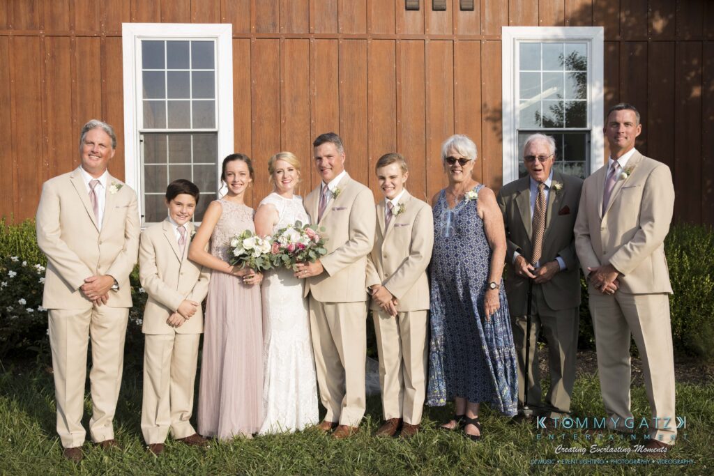 JenniferRudiWeddingFormalPhotos-214 - TOMMY GATZ ENTERTAINMENT A group of people in formal attire are standing together outdoors in front of a wooden building. The women hold bouquets.