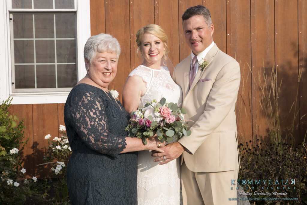 JenniferRudiWeddingFormalPhotos-217 - TOMMY GATZ ENTERTAINMENT A bride stands between an older woman in a dark dress and a man in a beige suit, holding a bouquet, in front of a wooden building.
