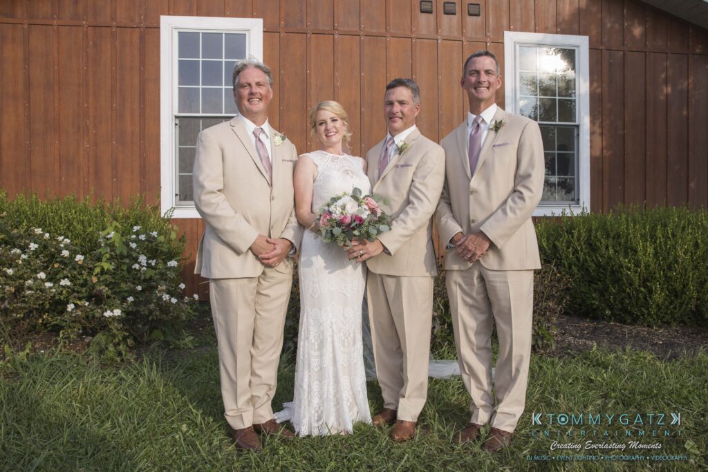 JenniferRudiWeddingFormalPhotos-220 - TOMMY GATZ ENTERTAINMENT Bride in a white lace dress and three men in beige suits stand together, smiling, in front of a wooden building with flowers and green grass.