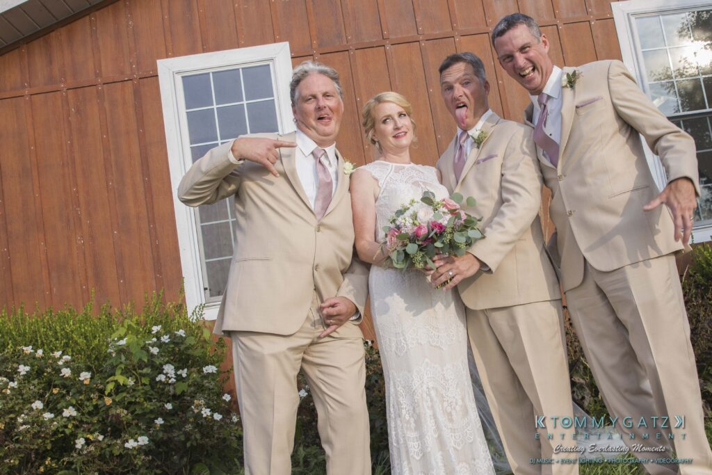 JenniferRudiWeddingFormalPhotos-222 - TOMMY GATZ ENTERTAINMENT A bride in a white dress stands with three men in beige suits making playful gestures in front of a wooden building with windows and nearby greenery.