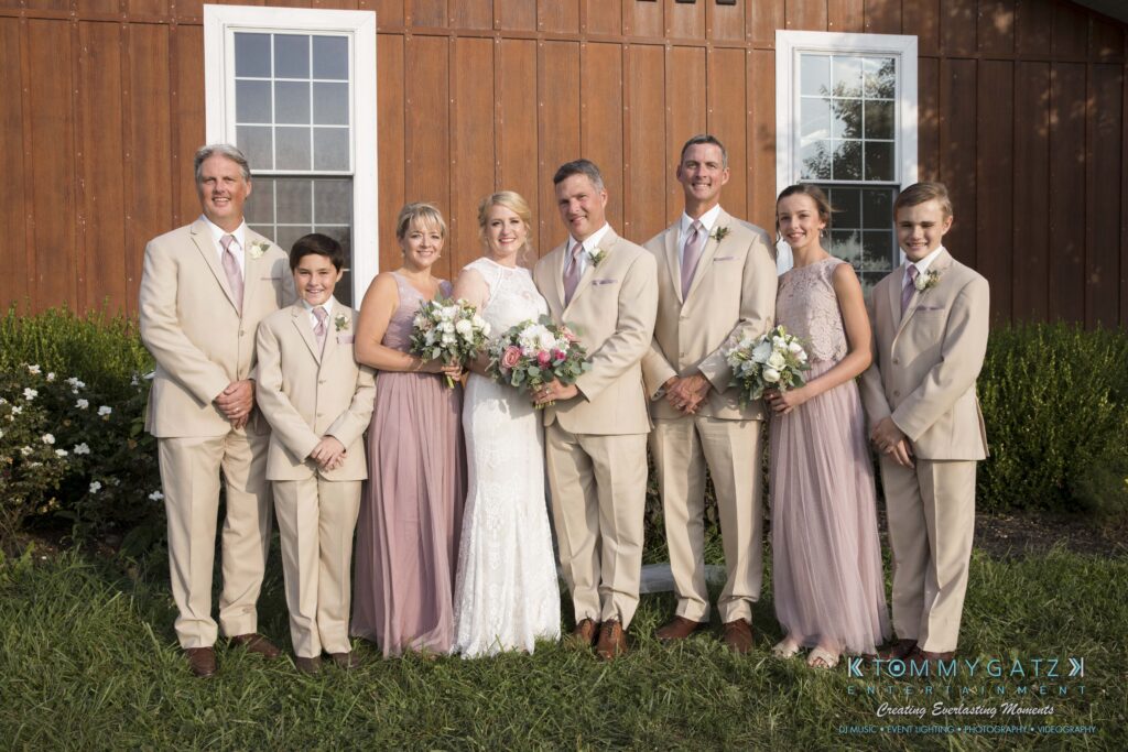 JenniferRudiWeddingFormalPhotos-224 - TOMMY GATZ ENTERTAINMENT A wedding group poses in front of a wooden building. The bride and groom are surrounded by six people, all dressed in beige and pink attire.