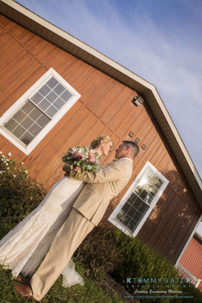 JenniferRudiWeddingFormalPhotos-229 - TOMMY GATZ ENTERTAINMENT Bride and groom in wedding attire stand closely facing each other, holding a bouquet, in front of a wooden building with green bushes and clear sky.