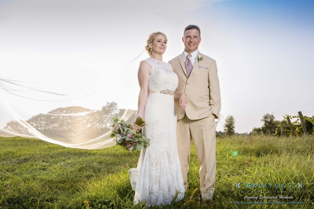 JenniferRudiWeddingFormalPhotos-233-1 - TOMMY GATZ ENTERTAINMENT A bride and groom in formal attire stand together outdoors in a grassy field. The bride holds a bouquet, and her veil is extended behind them.