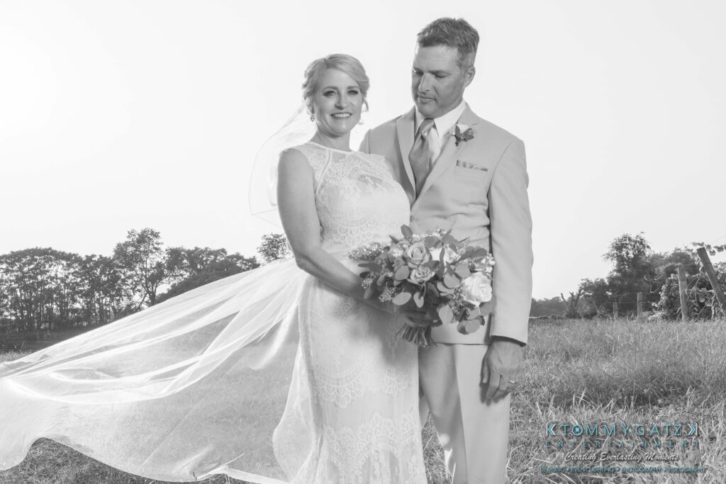 JenniferRudiWeddingFormalPhotos-234 - TOMMY GATZ ENTERTAINMENT Bride in a lace dress and groom in a suit stand outdoors. The bride holds a floral bouquet, and her veil flows in the breeze.