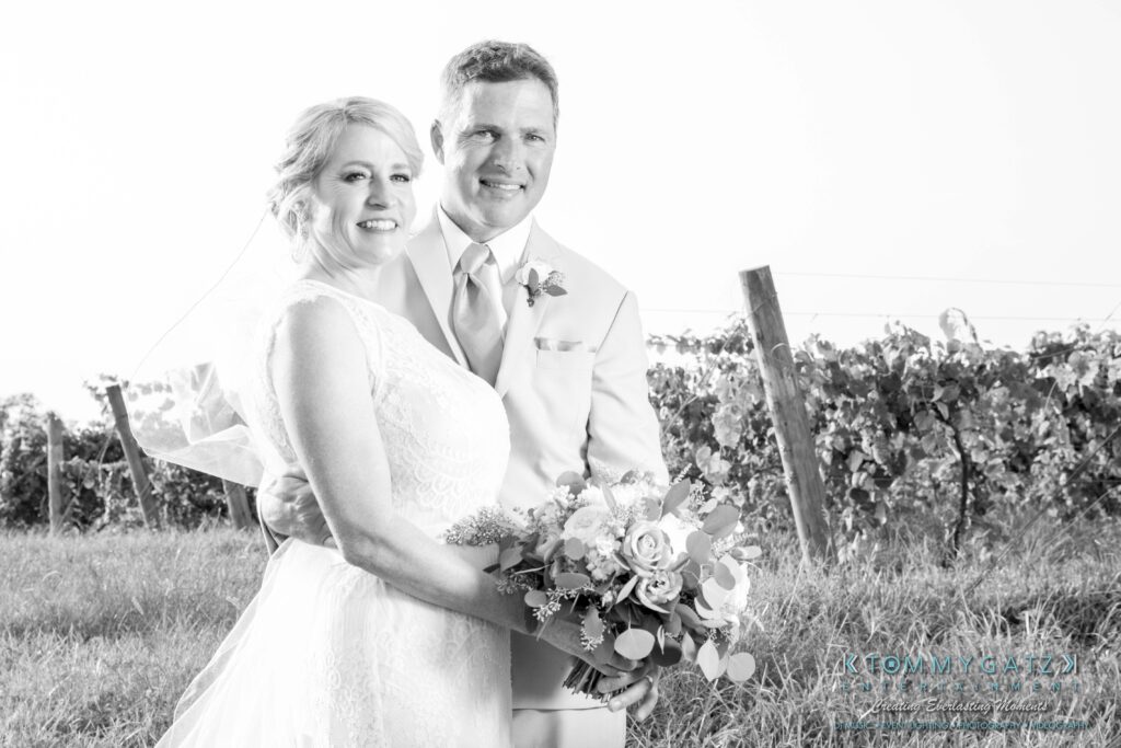 JenniferRudiWeddingFormalPhotos-236 - TOMMY GATZ ENTERTAINMENT A couple in wedding attire poses outdoors near a vineyard. The bride holds a bouquet, and both are smiling. The image is in black and white.