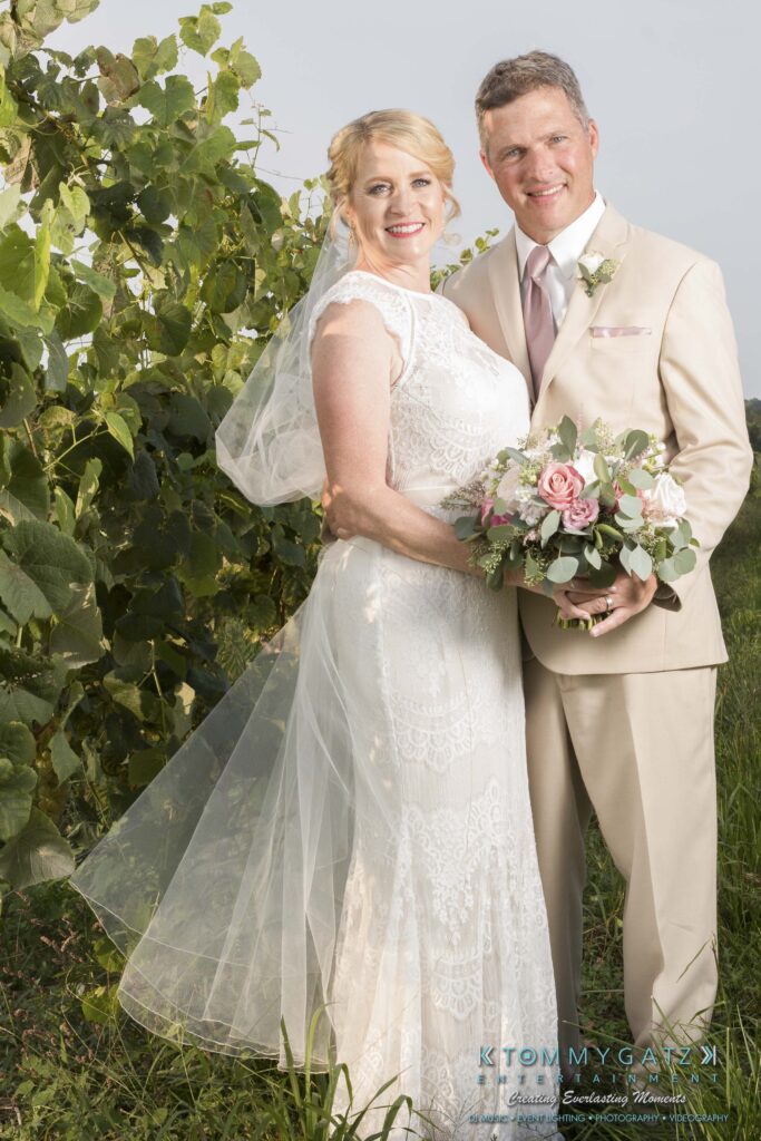 JenniferRudiWeddingFormalPhotos-237-1 - TOMMY GATZ ENTERTAINMENT A bride and groom in formal attire stand together in a garden setting, holding a bouquet of flowers.