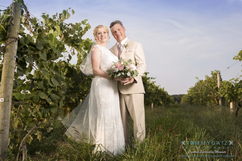 JenniferRudiWeddingFormalPhotos-239 - TOMMY GATZ ENTERTAINMENT A bride and groom stand together in a vineyard, with the bride holding a bouquet. They are both smiling, dressed in formal wedding attire.