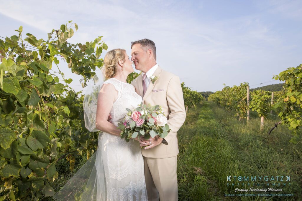 JenniferRudiWeddingFormalPhotos-244-1 - TOMMY GATZ ENTERTAINMENT A bride and groom embrace and smile at each other in a vineyard, with the bride holding a bouquet of pink and white flowers.
