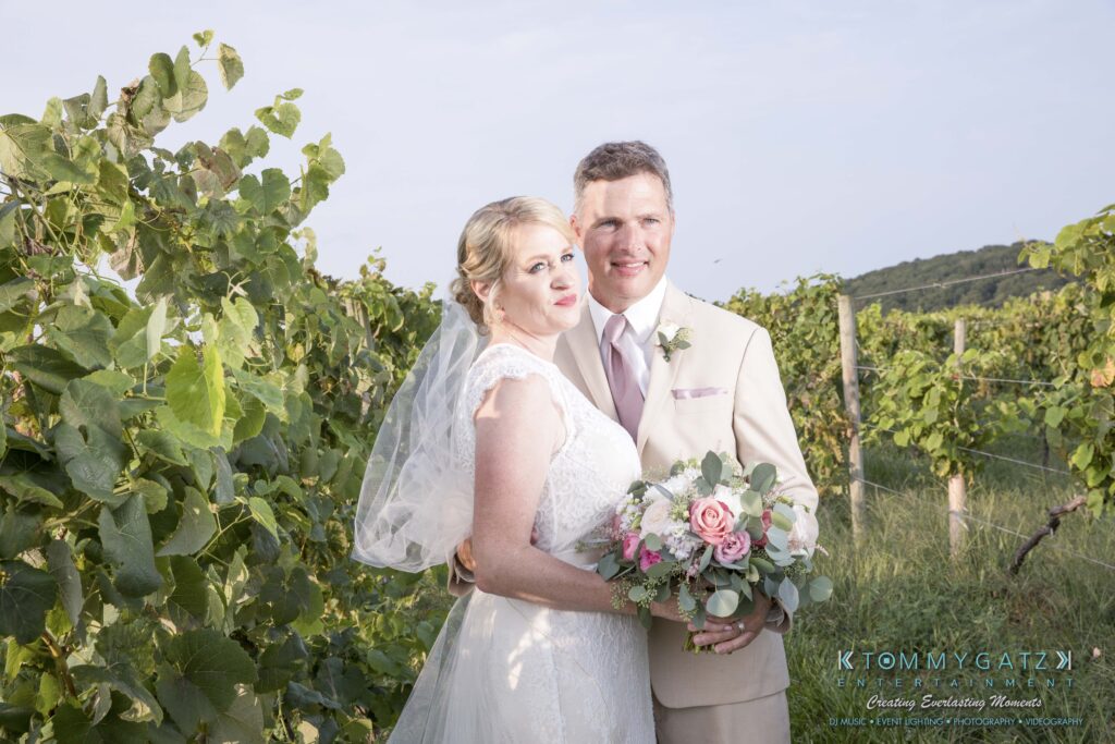 JenniferRudiWeddingFormalPhotos-245-1 - TOMMY GATZ ENTERTAINMENT Bride and groom pose together in a vineyard; the bride holds a bouquet.
