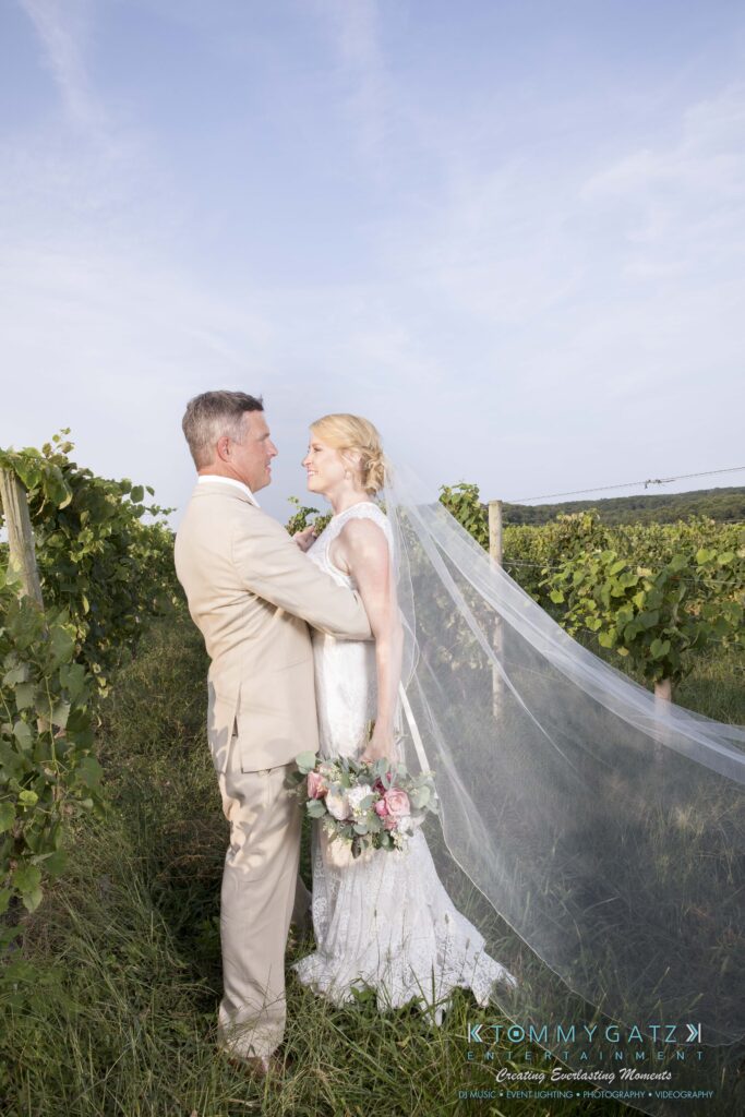 JenniferRudiWeddingFormalPhotos-249-1 - TOMMY GATZ ENTERTAINMENT A bride and groom stand together in a vineyard, with the bride wearing a long veil and holding a bouquet. Both are dressed in light-colored formal attire.