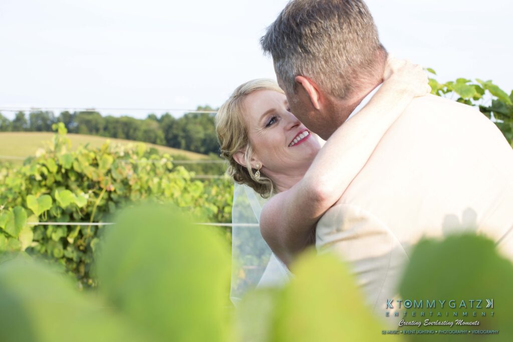 JenniferRudiWeddingFormalPhotos-251 - TOMMY GATZ ENTERTAINMENT A couple embraces in a vineyard. The woman smiles at the man, who has his back to the camera. Lush greenery surrounds them.