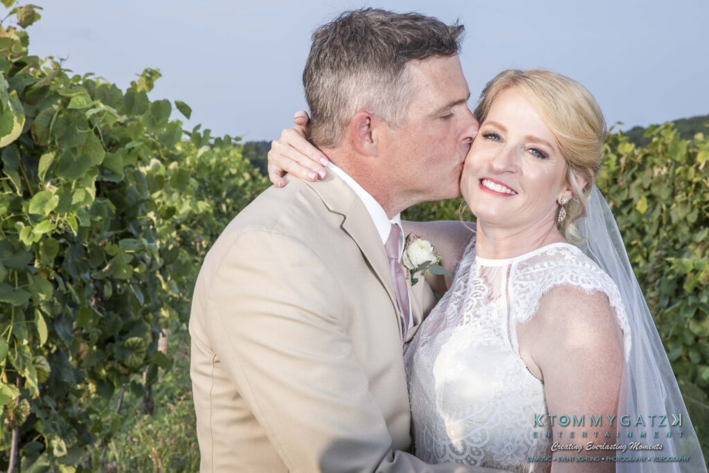 JenniferRudiWeddingFormalPhotos-253 - TOMMY GATZ ENTERTAINMENT A groom in a beige suit kisses a smiling bride in a white dress with a high neckline in a vineyard setting.