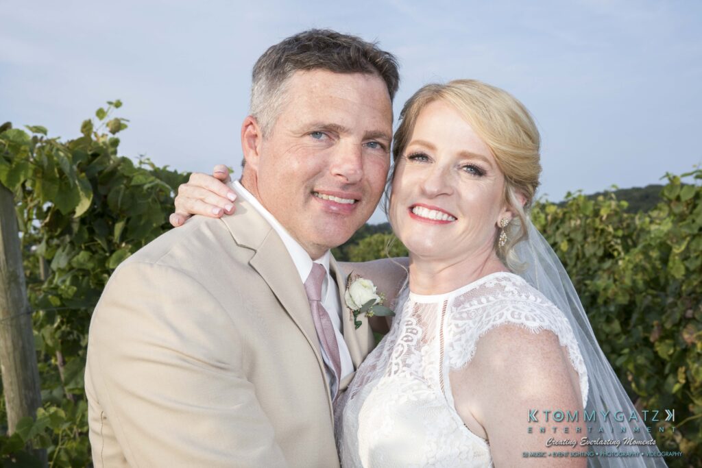 JenniferRudiWeddingFormalPhotos-254-1 - TOMMY GATZ ENTERTAINMENT A couple in wedding attire poses outdoors in a vineyard, smiling at the camera. The woman wears a white lace dress; the man wears a beige suit with a boutonniere.