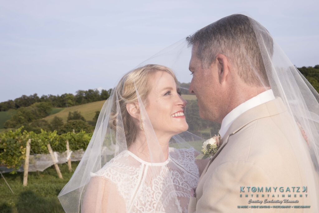 JenniferRudiWeddingFormalPhotos-256 - TOMMY GATZ ENTERTAINMENT Bride and groom smiling at each other under a wedding veil in an outdoor setting with greenery in the background.