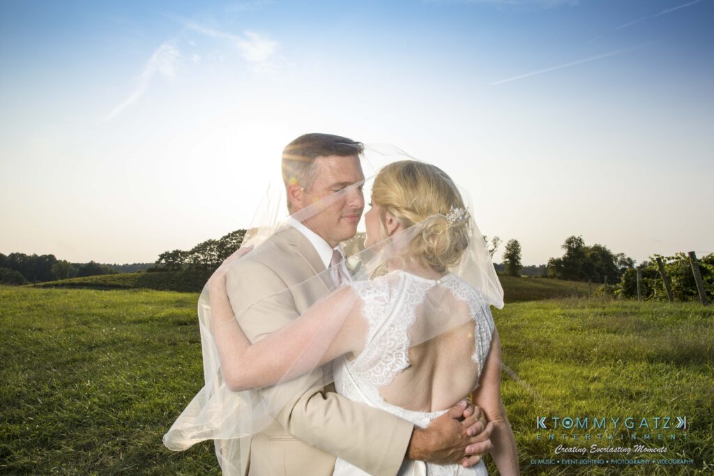 JenniferRudiWeddingFormalPhotos-257 - TOMMY GATZ ENTERTAINMENT A bride and groom embrace outdoors in a grassy field under a clear sky. The bride's veil is draped over both of them.