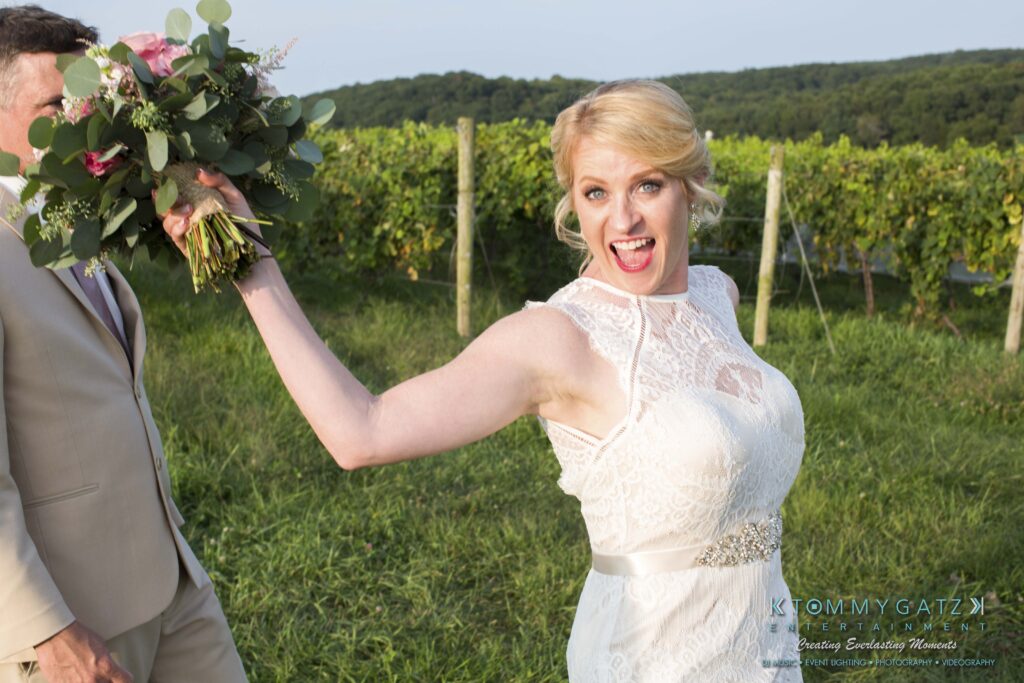 JenniferRudiWeddingFormalPhotos-258 - TOMMY GATZ ENTERTAINMENT Bride in a white dress energetically holds up a bouquet, smiling broadly, with greenery in the background.
