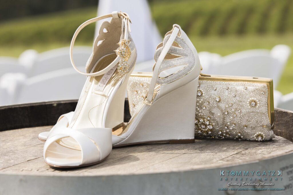 JenniferRudiWeddingPrepPhotos-10 - TOMMY GATZ ENTERTAINMENT White wedge sandals with decorative ankle straps are placed on a wooden surface next to a gold-embellished clutch.