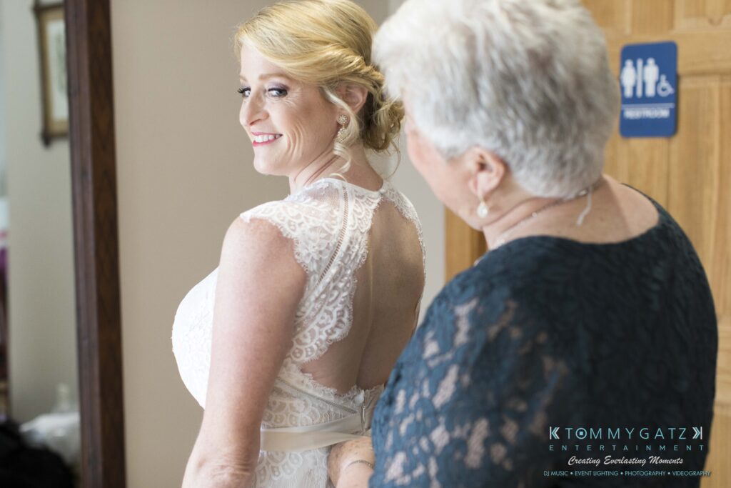 JenniferRudiWeddingPrepPhotos-37 - TOMMY GATZ ENTERTAINMENT A woman in a white lace dress looks back and smiles as another woman helps her adjust the dress in a room.