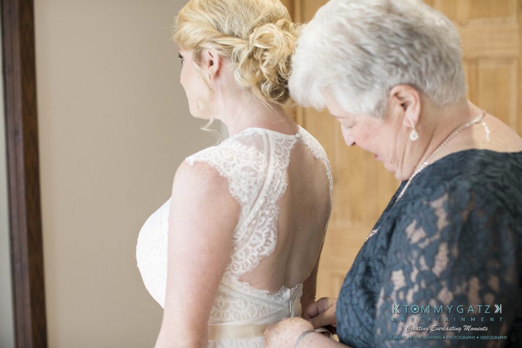 JenniferRudiWeddingPrepPhotos-38 - TOMMY GATZ ENTERTAINMENT A woman in a lace wedding dress is being helped with her dress by another woman in a dark outfit.