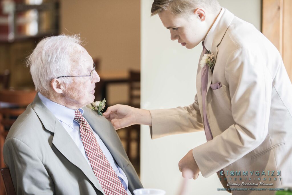 JenniferRudiWeddingPrepPhotos-48 - TOMMY GATZ ENTERTAINMENT A young man adjusts an elderly man's boutonniere. Both are in suits, in a well-lit room with chairs in the background.