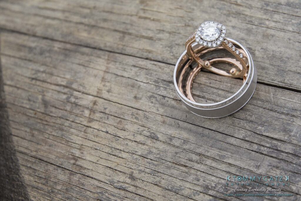 JenniferRudiWeddingPrepPhotos-7 - TOMMY GATZ ENTERTAINMENT A set of wedding rings, featuring a diamond engagement ring and a silver band, rests on a wooden surface.