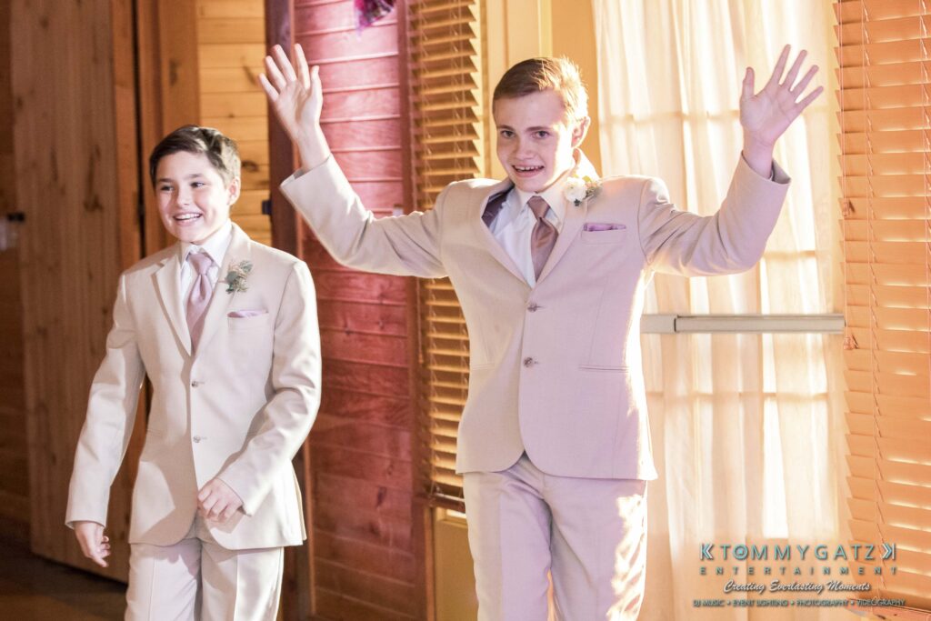 JenniferRudiWeddingReceptionPhotos-294 - TOMMY GATZ ENTERTAINMENT Two boys in beige suits enter a room, the boy on the right raising his arms, both smiling.