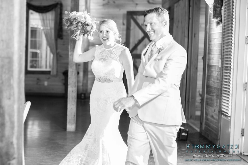 JenniferRudiWeddingReceptionPhotos-296 - TOMMY GATZ ENTERTAINMENT Bride and groom in wedding attire enter a venue holding hands. Bride holds a bouquet. Black and white photo.