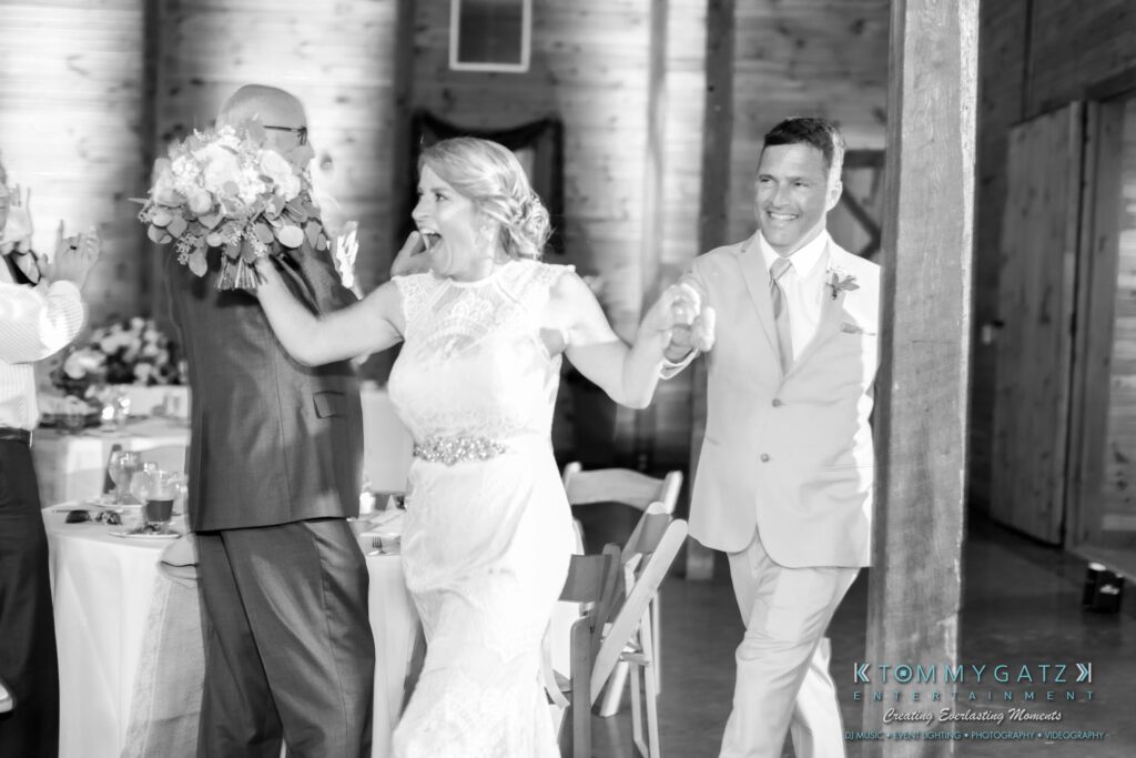 JenniferRudiWeddingReceptionPhotos-297 - TOMMY GATZ ENTERTAINMENT Bride and groom joyfully enter a reception venue holding hands. The bride holds a bouquet and wears a lace dress, while the groom is in a light-colored suit.