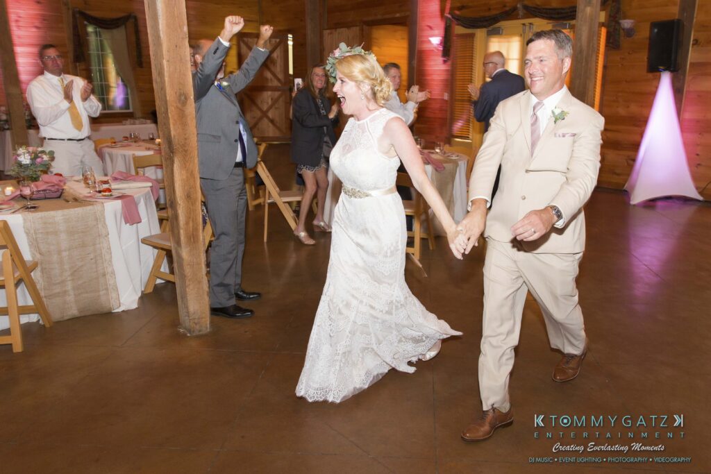 JenniferRudiWeddingReceptionPhotos-298 - TOMMY GATZ ENTERTAINMENT A joyful bride and groom enter a rustic reception hall, where guests cheer and celebrate their arrival.