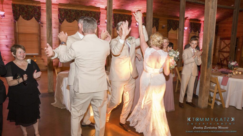 JenniferRudiWeddingReceptionPhotos-299 - TOMMY GATZ ENTERTAINMENT A bride and groom dance in a warmly lit venue, surrounded by clapping guests. The couple is dressed in light formal wear, and the bride holds a bouquet. Tables are set in the background.