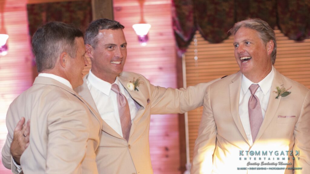 JenniferRudiWeddingReceptionPhotos-300 - TOMMY GATZ ENTERTAINMENT Three men in beige suits and ties are smiling and conversing inside a warmly lit room. A logo with "Tommy Gatz Entertainment" is visible at the bottom.