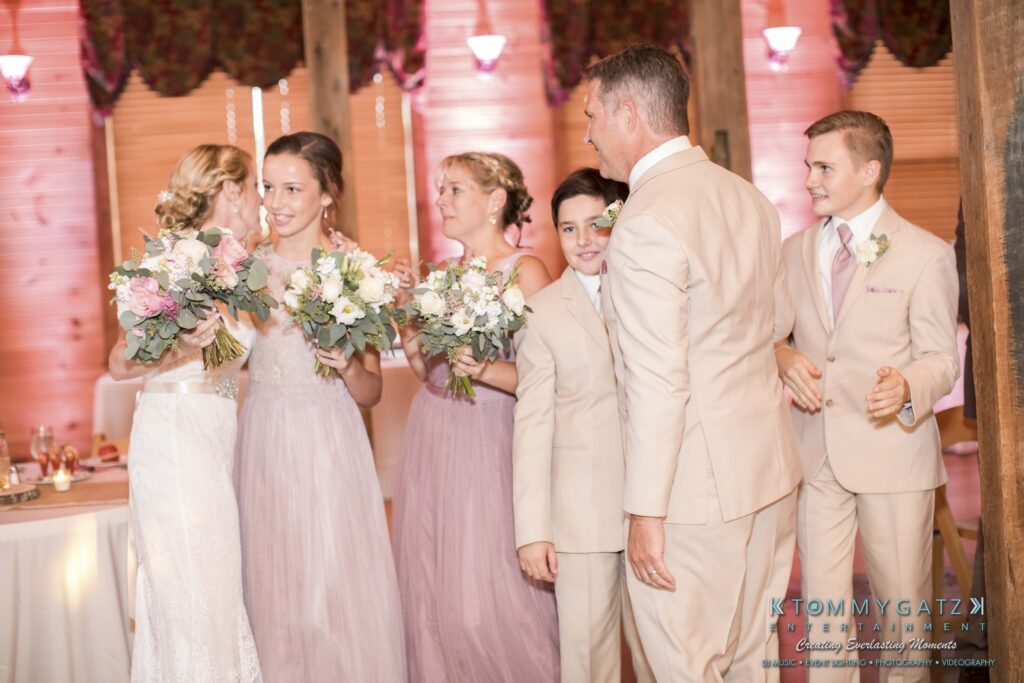 JenniferRudiWeddingReceptionPhotos-304 - TOMMY GATZ ENTERTAINMENT A wedding party in light attire stands indoors. Three women hold floral bouquets, while three men watch. A decorated table is visible in the background.