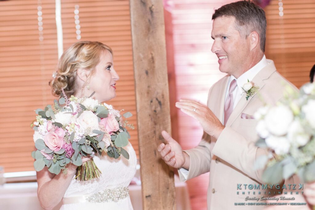 JenniferRudiWeddingReceptionPhotos-308 - TOMMY GATZ ENTERTAINMENT A bride holding a bouquet converses with a groom in a light suit at an indoor venue.