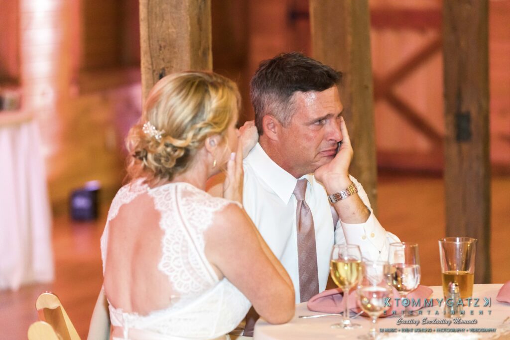 JenniferRudiWeddingReceptionPhotos-317 - TOMMY GATZ ENTERTAINMENT A couple sits at a table during a formal event. The woman has her back turned slightly, while the man looks contemplative. Glasses and dishes are on the table; a wooden structure is behind them.
