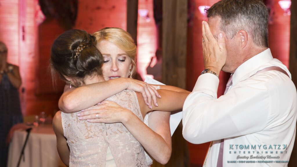 JenniferRudiWeddingReceptionPhotos-323 - TOMMY GATZ ENTERTAINMENT Two women embrace in a hug at a gathering while a man nearby wipes his eyes. A sign in the foreground reads "Tommy Gatz Entertainment.