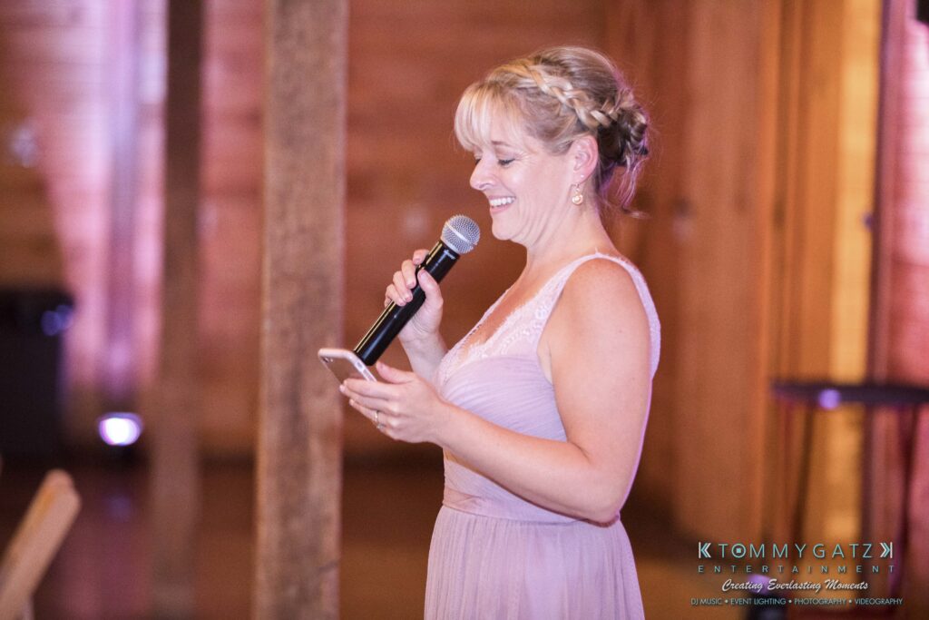 JenniferRudiWeddingReceptionPhotos-326 - TOMMY GATZ ENTERTAINMENT Woman in a lavender dress holds a microphone and a phone, standing in a wooden venue.