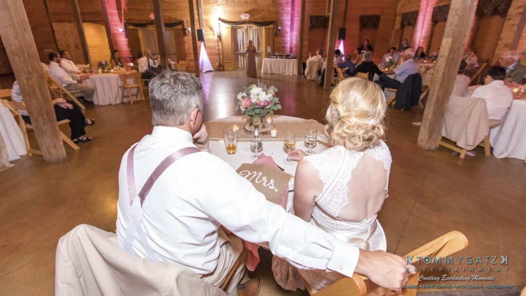 JenniferRudiWeddingReceptionPhotos-327 - TOMMY GATZ ENTERTAINMENT A couple sits at a wedding reception table, facing the dance floor. A "Mrs." sign is visible on the chair.