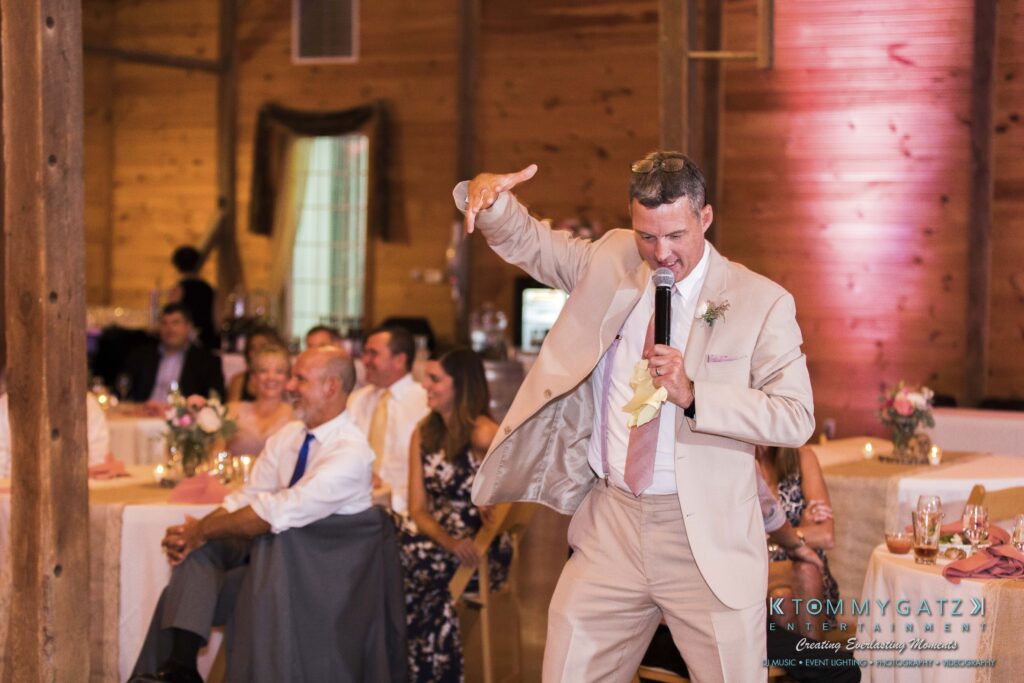 JenniferRudiWeddingReceptionPhotos-332 - TOMMY GATZ ENTERTAINMENT A man in a suit and tie is speaking into a microphone at an indoor event. Seated guests are watching him. The setting is decorated with flowers and has wooden walls.