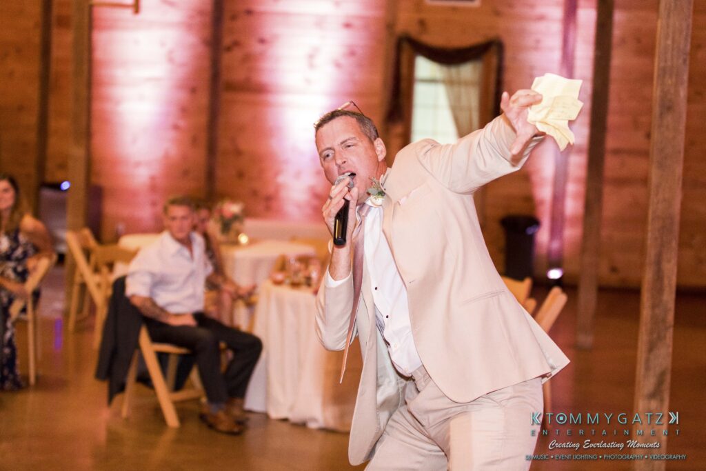 JenniferRudiWeddingReceptionPhotos-333 - TOMMY GATZ ENTERTAINMENT A man in a light suit holds a microphone and paper, speaking energetically in a decorated venue, with guests seated at tables in the background.